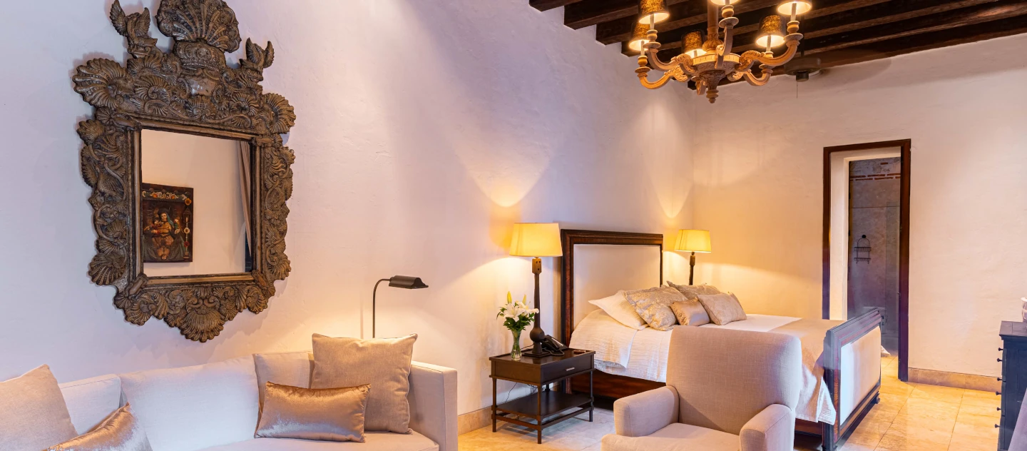 Dormitorio elegante con iluminación cálida, espejo tallado artesanalmente, techo de vigas oscuras y sala de estar integrada