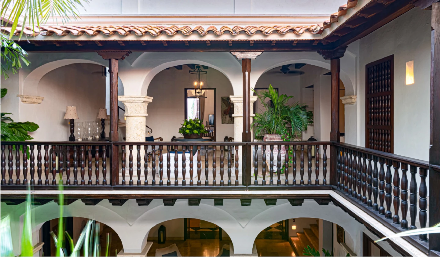 Pasillo superior de una casa colonial de lujo con arcos de medio punto, barandillas de madera tallada y un patio interior luminoso decorado con plantas tropicales.